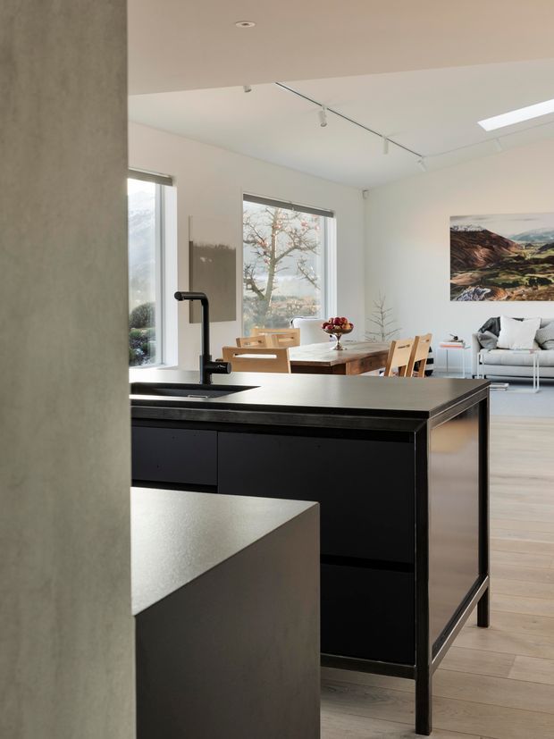 A bright, open-plan contemporary kitchen viewed from behind a partial wall, featuring a large matte-black island with an integrated sink and tall black tap. The kitchen opens to a dining area with a timber dining table and chairs and a light-coloured living room beyond. Pale timber flooring, white walls, large windows, a skylight, track lighting, and mountain artwork create an airy, minimal interior.
