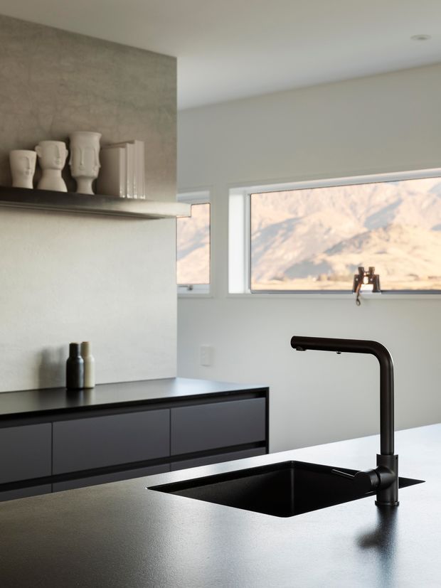 A minimalist contemporary kitchen focused on a dark matte island benchtop with an integrated black undermount sink and tall black gooseneck tap. Dark flat-panel cabinetry lines the rear wall, beneath a concrete-look feature wall and open shelf displaying sculptural ceramic vessels. Large horizontal windows bring in natural light and frame a rocky landscape.