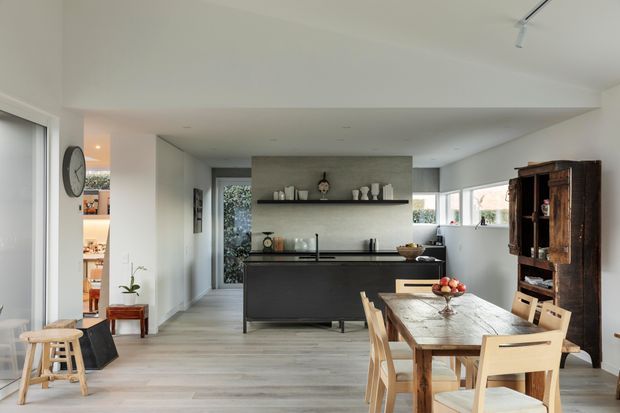 A bright open-plan kitchen and dining area with a contemporary dark kitchen island, pale timber flooring, white walls, and broad windows. A long timber dining table with light wood chairs occupies the foreground, while a tall rustic timber display cabinet provides contrast against the clean-lined kitchen. A small pantry or utility area is visible through the opening on the left.