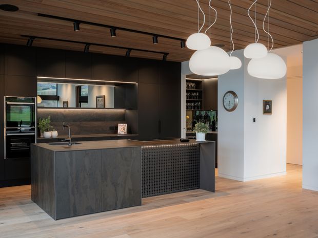 A sophisticated open-plan kitchen with a large dark island, integrated black cabinetry, a textured feature front, and a timber-lined ceiling. Warm timber flooring and ceiling boards contrast with the charcoal joinery, while sculptural white pendant lights hang above the island. A dining or living area is partially visible beyond the kitchen.