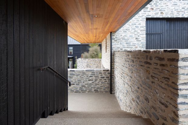 A covered exterior passageway with rough pale stone masonry walls, dark vertical timber cladding and doors, and a warm timber-lined soffit with recessed lighting. The textured concrete or stone-paved walkway slopes gently through the architectural composition, creating a sheltered, contemporary courtyard-like circulation space.
