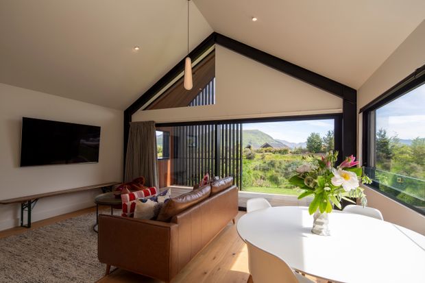 Bright contemporary open-plan living and dining area with a vaulted ceiling, exposed dark structural framing, extensive floor-to-ceiling glazing, and views toward a landscaped rural setting and mountains. A brown leather sofa faces a wall-mounted television, while a white rounded dining table sits beside the windows. Timber flooring, neutral walls, vertical screen elements, and a pendant light create a warm, refined interior.