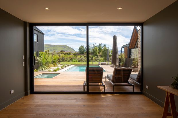 A minimalist interior room with dark painted walls, recessed ceiling lights, and a warm timber floor opening through full-height sliding glass doors to a timber deck, swimming pool, landscaped garden, and lounge area. Hills and neighboring buildings are visible beyond the outdoor space.