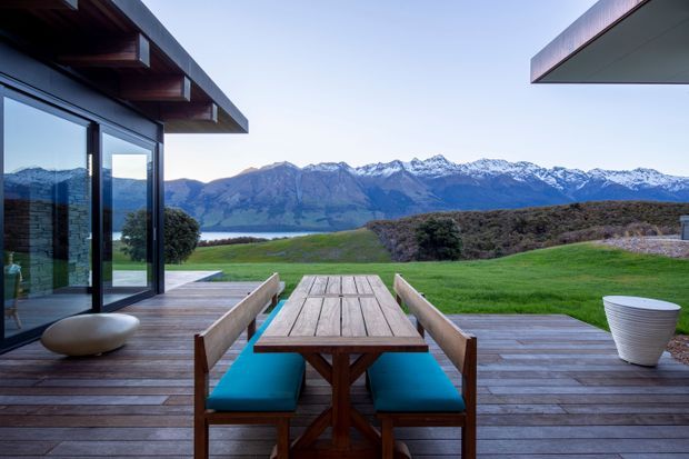 A contemporary lakeside outdoor dining terrace overlooking a broad lake and snow-capped mountain range. The deck uses warm timber boards and includes a timber dining table with built-in bench seating and vivid turquoise cushions. Floor-to-ceiling dark-framed glazing connects the terrace to the modern home, with broad roof overhangs and a neatly landscaped lawn beyond.