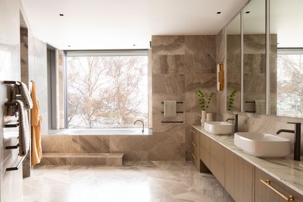 Bright, spacious contemporary bathroom with a freestanding bathtub positioned by a large window, a double vanity with vessel basins, mirrored cabinetry, wall-mounted towel rails, and warm stone-look wall and floor finishes. Natural light fills the room and a leafy outdoor view is visible through the glazing.
