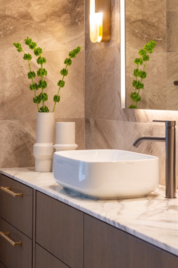 Close-up of an elegant bathroom vanity featuring a white vessel basin, tall chrome tapware, dark timber-toned cabinetry with brass pulls, and a white marble countertop. Large beige-brown stone-look wall tiles surround the vanity, with a backlit mirror and warm wall sconce above. Decorative green stems in neutral ceramic vessels add a natural accent.