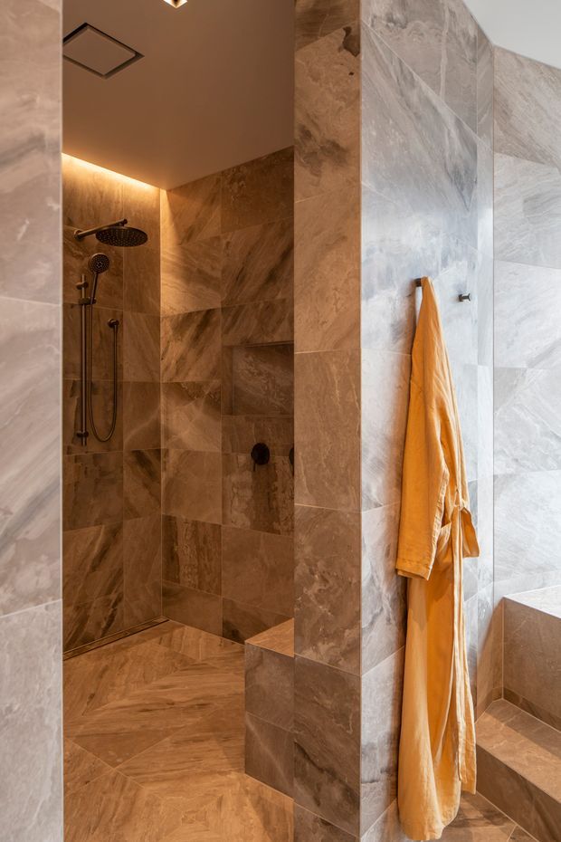 A warm, luxurious bathroom featuring a walk-in shower with a rain showerhead and handheld fixture. The shower, walls, floor, and built-in bath surround are clad in large-format veined stone-look tiles in brown, taupe, and grey tones. A partial freestanding or built-in bathtub is visible on the right, with a vivid orange towel adding contrast. Recessed ceiling lighting and concealed linear lighting create a softly illuminated atmosphere.