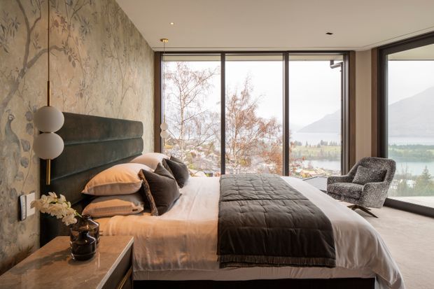 A refined bedroom with a king-sized bed positioned beside expansive floor-to-ceiling glazing. The room features a tall upholstered dark green headboard, layered neutral and charcoal bedding, a marble-topped bedside table, sculptural spherical pendant lighting, patterned botanical wallcovering, and a textured lounge chair. Trees, a lake, and mountains are visible outside, contributing to a serene, nature-connected atmosphere.