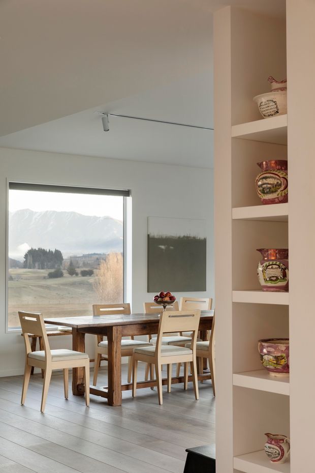 A bright dining area viewed from an adjacent space, with a large rustic timber dining table surrounded by light upholstered chairs. A broad window frames a rural mountain landscape, while a built-in open shelving unit displays decorative ceramics along the right side. The room has pale walls, light grey timber flooring, a sloping ceiling, and understated contemporary detailing.