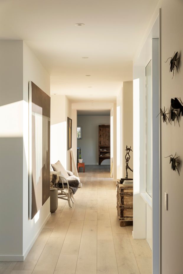 A bright, elongated residential hallway with warm pale timber floorboards, white walls, recessed ceiling lights, and strong sunlight entering from the side. A seating nook with a chair and cushions sits along the left wall, while rustic timber storage and sculptural decorative pieces line the right. Artwork and insect-shaped wall ornaments add visual interest, with a doorway and timber cabinet visible at the far end.