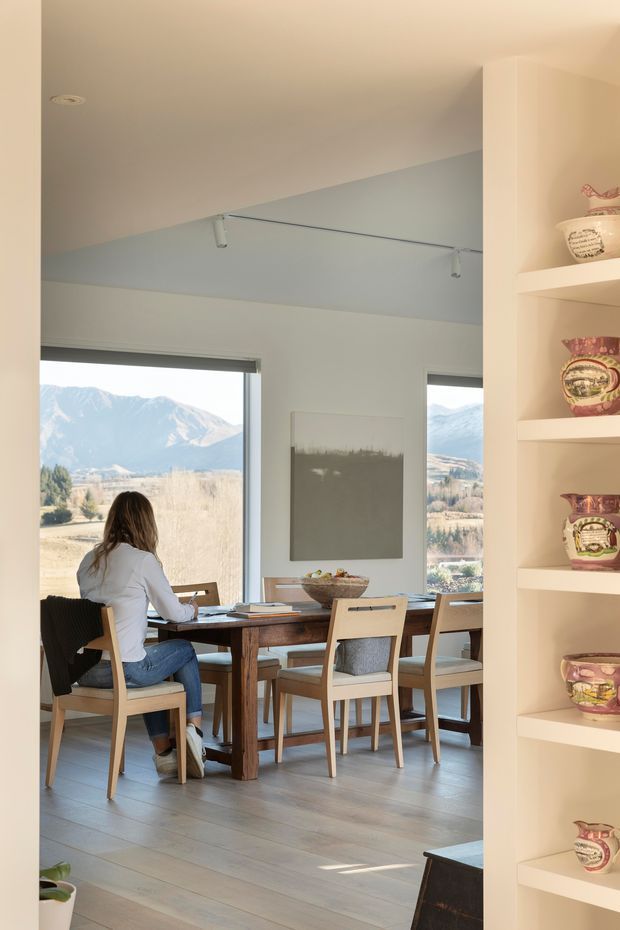 Bright dining area viewed from an adjoining space, with a timber dining table and chairs positioned beside large windows overlooking a mountainous landscape. The room has pale walls, light timber flooring, simple track lighting, a neutral artwork panel, and built-in cream shelving displaying decorative ceramics. A person is seated at the table, and the space feels airy and warm.