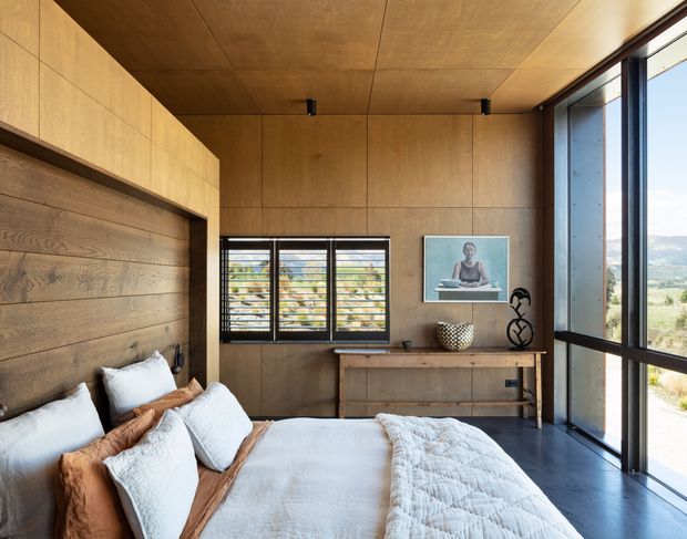 A warm, compact bedroom clad in timber panels, with a built-in timber bedhead and integrated storage along the left wall. The room has a low timber-lined ceiling, dark flooring, a small horizontal louvred window, and expansive dark-framed glazing on the right overlooking a rural landscape. Bedding and cushions create a soft, relaxed focal point, complemented by a small timber dresser and understated artwork.
