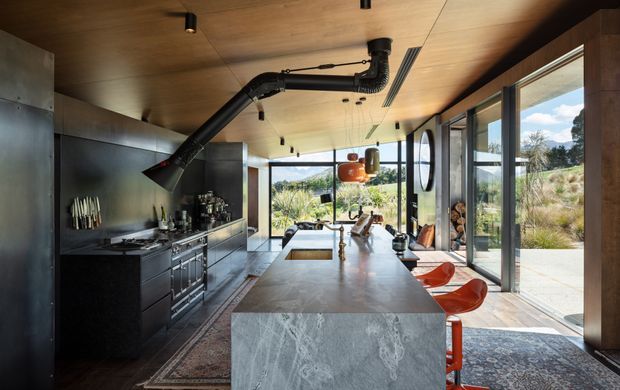 A sophisticated open-plan kitchen with a large stone island, dark metal cabinetry, exposed black extraction ductwork, and a warm timber ceiling. Full-height glazing opens to a landscaped outdoor setting, while sculptural orange bar stools and pendant lighting add colour. The kitchen has a refined industrial character with strong indoor-outdoor connection.