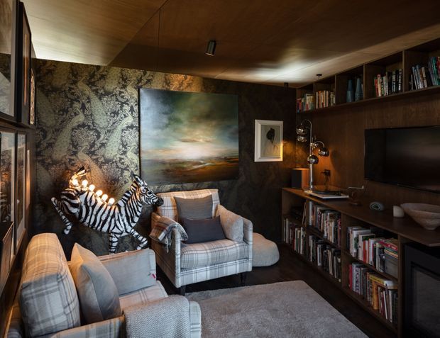 A compact, richly layered living room or media room with dark timber wall and ceiling panelling, extensive built-in bookshelves, a wall-mounted television, and a fireplace at right. Two large plaid armchairs and additional seating face into the room, while a zebra sculpture, patterned feature wallpaper, framed artwork, and sculptural lighting add eclectic character. The space is warm, dimly lit, intimate, and highly curated.
