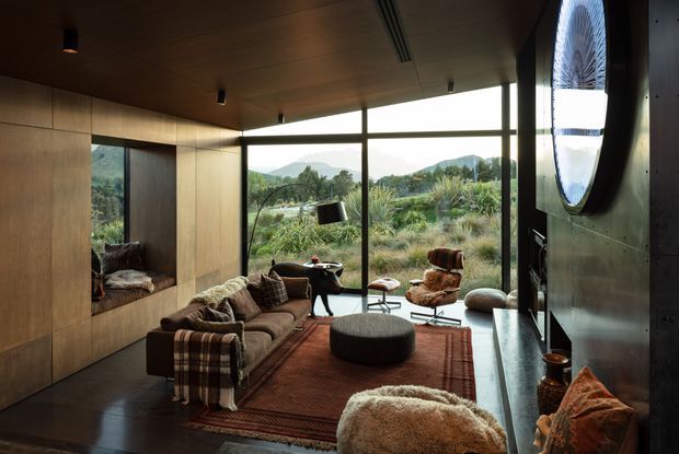 A warm, sophisticated living room with extensive floor-to-ceiling glazing overlooking a landscaped natural setting and distant hills. The room features richly toned timber wall and ceiling lining, dark flooring, a large brown sofa with patterned throws, a rust-toned rug, sculptural lounge chairs, a low round ottoman, and a dramatic oval window detail. The interior has a refined mid-century-inspired character with strong natural materials and a connection to the outdoors.