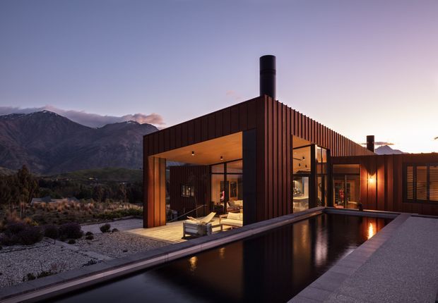 A contemporary single-storey residence at dusk, clad in dark vertical timber or metal-like slats, with extensive floor-to-ceiling glazing opening onto a paved terrace and long reflecting pool. Warm interior and exterior lighting contrasts with the dark facade, while rugged mountains form the distant backdrop.