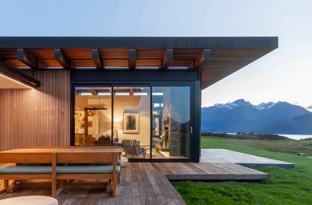 Contemporary single-storey pavilion-like home with extensive glazing opening onto a timber deck and outdoor dining area. The dark flat roof has exposed timber soffit framing and deep eaves, while warm vertical timber cladding and decking contrast with the black-framed sliding glass doors. A bright living area is visible inside, with a dramatic mountain and lakeside landscape beyond the property.