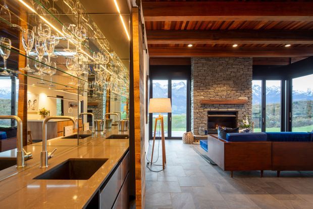 Open-plan luxury kitchen and living room with a long stone island, dark integrated cabinetry, mirrored glass shelving displaying glassware, and multiple sinks and taps. The adjacent living area has a blue sofa, timber-lined ceiling with exposed beams, a substantial stacked-stone fireplace, and expansive glazing overlooking a mountainous landscape. The interior combines warm timber, stone, dark metal framing, and polished surfaces.