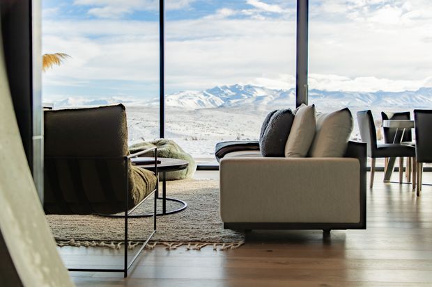 A refined living room with large floor-to-ceiling glazing overlooking a dramatic snow-covered mountain landscape. The room features a neutral upholstered sofa and armchair, slim black metal side tables, a textured area rug, and warm timber flooring. A dining area is visible at the right, while the expansive glazing creates a strong connection to the alpine setting.