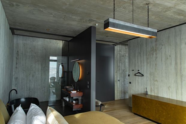 A moody, industrial-style bedroom with a concrete-look ceiling and vertically textured grey wall finishes. The room includes a partially visible bed with white pillows, dark leather seating, a black open vanity or washstand with round mirrors, timber flooring, a mustard-gold built-in cabinet, and a suspended rectangular pendant light. Black-framed glazing and dark partitions add strong graphic contrast.