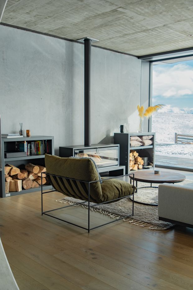 A modern living room with a minimalist black wood-burning stove centered against a raw concrete wall, with a large corner window overlooking a snowy mountain landscape. The room features warm timber flooring, an open black metal-framed firewood storage unit, a low timber-and-black coffee table, a textured neutral rug, and a dark olive lounge chair. The exposed concrete ceiling and walls, restrained furnishings, and expansive glazing create a refined alpine-industrial atmosphere.