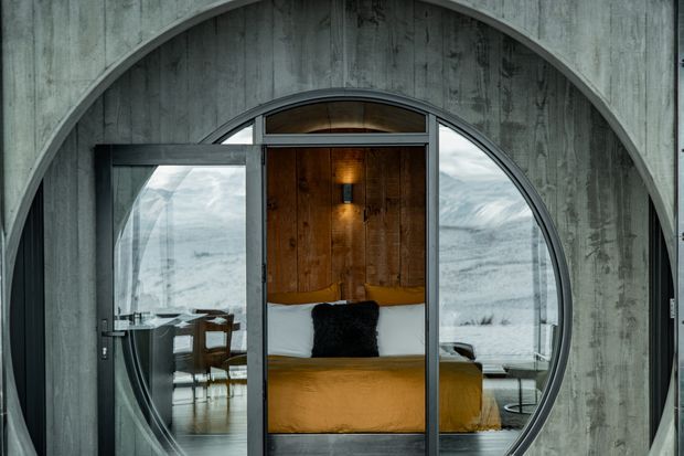 A dramatic architectural interior viewed through a large circular glazed opening set into a textured grey concrete wall. The central opening frames a compact bedroom with warm timber wall panelling, a wall light, white pillows, a dark accent cushion and ochre bedding. Reflective glazing on either side reveals a dining or living area with timber furniture and expansive mountainous scenery beyond. The composition contrasts cool concrete and glass with warm timber and textiles.