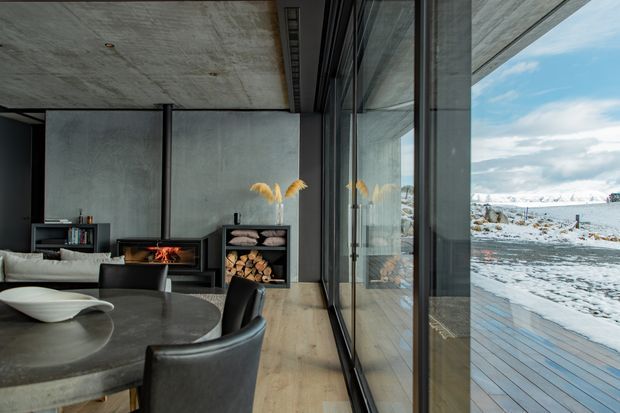 A contemporary open-plan living and dining area with a polished dark stone dining table, upholstered dining chairs, a low light-coloured sofa, black built-in shelving and a fireplace with stacked firewood. Full-height glazing runs along one side, reflecting the interior and overlooking a snow-covered landscape and timber deck. The space features a raw concrete ceiling and grey concrete-look wall surfaces, light timber flooring, and a restrained charcoal, grey and neutral palette.