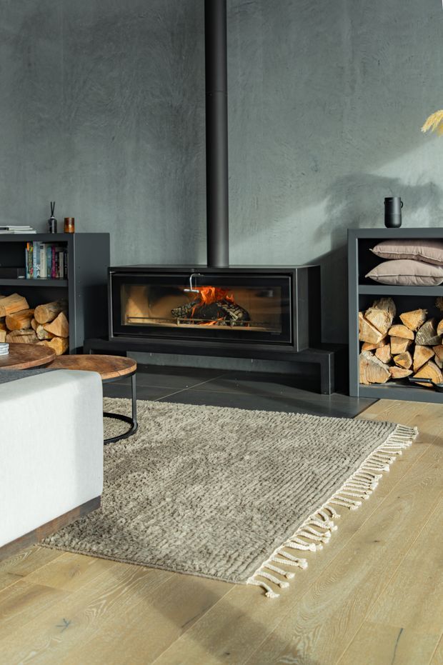 A cozy contemporary living room centered on a built-in black fireplace with a visible wood fire and black flue. The fireplace is set against a textured grey feature wall and flanked by dark open shelving storing books, cushions, and firewood. A pale upholstered armchair, round timber coffee tables, and a large textured fringed rug sit over light timber flooring. Warm sunlight and the fire contrast with the dark cabinetry and charcoal wall.