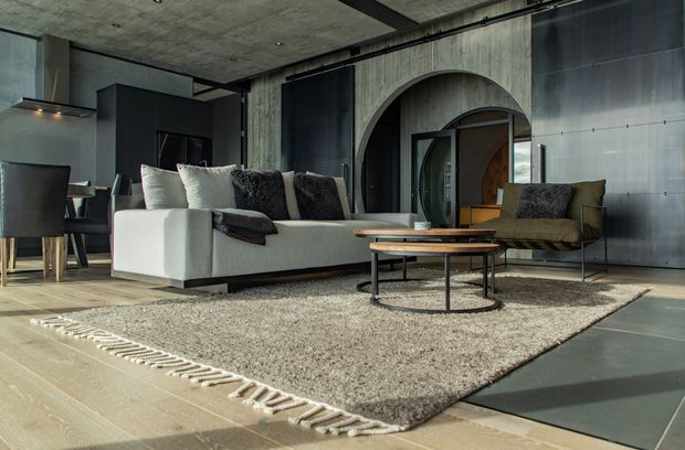 A sophisticated open-plan living room with a lounge and dining area. The space features a large pale sectional sofa, dark textured cushions, an olive accent armchair, layered round coffee tables, and a large neutral fringed rug. Raw concrete walls and ceiling, dark built-in cabinetry, exposed structural detailing, arched wall openings, and metallic wall cladding create a dramatic industrial character. Timber flooring and warm sunlight soften the otherwise cool, moody palette.