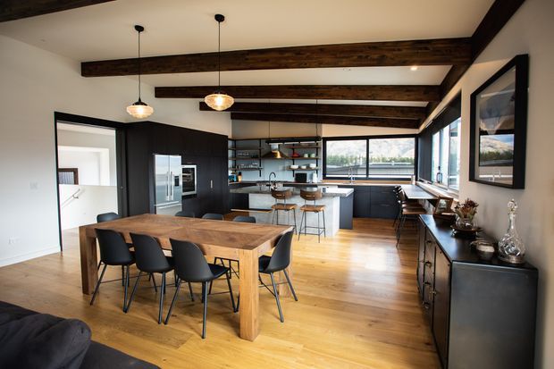 Open-plan dining and kitchen interior with a large solid timber dining table, black dining chairs, dark built-in cabinetry, a central island, exposed dark timber ceiling beams, pendant lights, timber flooring, and large windows.