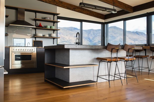 A contemporary kitchen with a large dark-and-grey island, integrated seating ledge, open timber shelving, stainless steel range cooker, and a row of bar stools. Floor-to-ceiling corner glazing provides expansive mountain views. Exposed dark timber ceiling beams and warm timber flooring add contrast to the minimalist cabinetry and concrete-look surfaces.