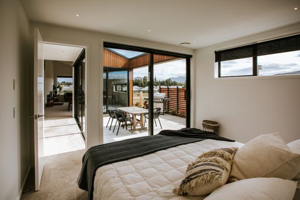 A contemporary bedroom with a neatly made bed, neutral bedding, carpeted floor and recessed ceiling lights. Large sliding glass doors open to a private outdoor terrace, with a horizontal window providing views toward the surrounding landscape. An adjacent hallway and part of another room are visible through the open doorway.
