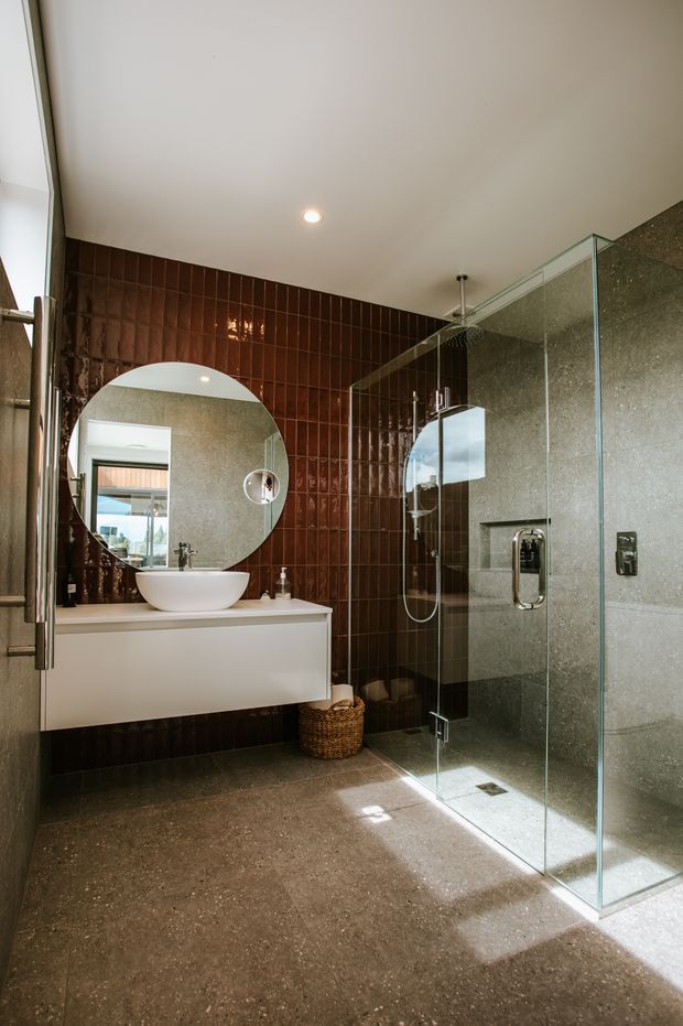 A contemporary bathroom featuring a floating white vanity with a vessel basin, a large round mirror, a frameless glass walk-in shower, dark glossy brown vertical wall tiles, and grey textured stone-look wall and floor finishes. Natural light enters from the left, creating a warm, refined atmosphere.