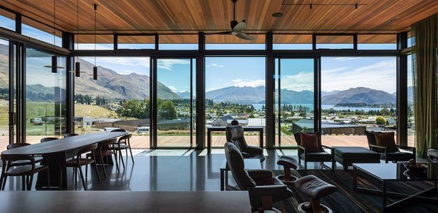 A spacious, glass-walled living and dining area with expansive views toward a mountainous lake or valley landscape. The room features a richly timber-lined ceiling, dark-framed floor-to-ceiling glazing, polished dark flooring, a long dining table with chairs, and a lounge grouping with leather seating and low tables. The overall design is refined, warm, and strongly connected to the landscape.