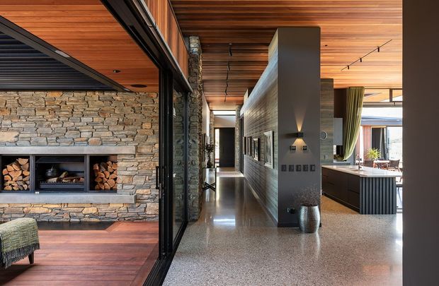 A refined open-plan residential interior with a long central hallway connecting a living area and kitchen. The living zone features a substantial stacked natural-stone fireplace and timber flooring, while the hallway and kitchen use polished terrazzo flooring, dark built-in joinery, and extensive warm timber ceiling lining. Black-framed glazed partitions create visual separation while maintaining openness. The kitchen includes a large island, and the space is lit by recessed and track lighting.