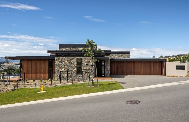 Street-facing view of a low, single-storey contemporary residence with a broad horizontal composition and low-pitched roof. The facade combines stacked natural stone around the entry with warm vertical timber garage and screening panels, dark window and door frames, and a large integrated double garage. A paved driveway, front lawn, young tree, and landscaped entry sit between the house and the suburban street.