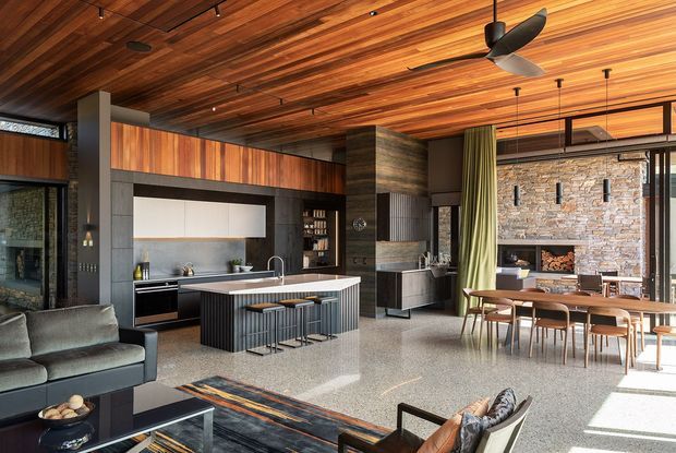 Open-plan contemporary living, kitchen and dining space with a dramatic exposed timber-lined ceiling, dark built-in cabinetry, a stone kitchen island with bar seating, and a dining area beside expansive glazing. A textured natural-stone fireplace wall forms a strong focal point, while polished terrazzo-like flooring, black-framed windows, and layered timber joinery create a warm, refined interior.
