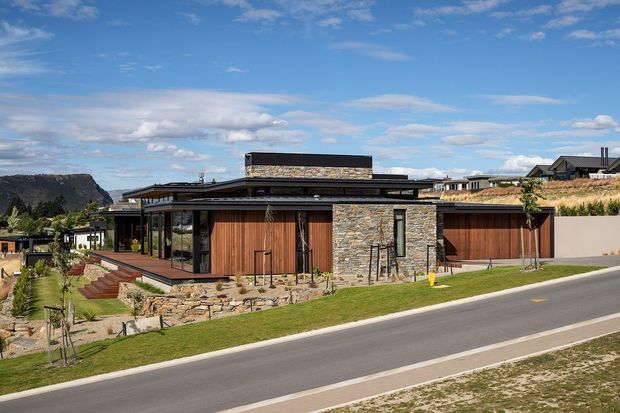 Exterior street view of a low, contemporary single-storey residence with broad flat roof planes, extensive glazing, natural stone walls, warm timber garage and entry doors, landscaped terraces, and a dry mountainous setting in the background.