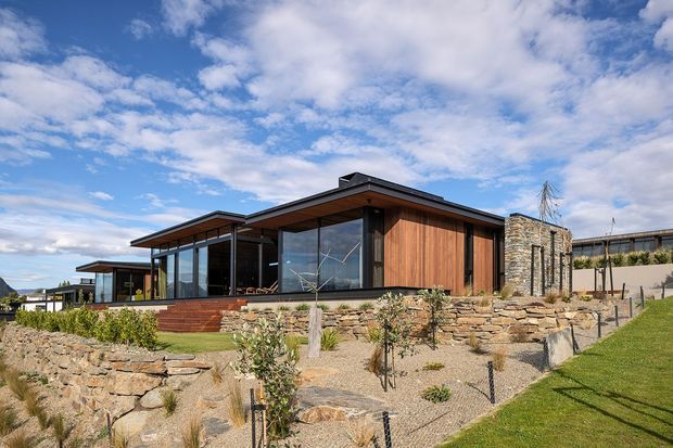 Single-storey contemporary house set on a landscaped, gently sloping site. The low-profile building has broad flat or very low-pitched roof planes, extensive floor-to-ceiling glazing, warm vertical timber wall cladding, dark metal framing, and substantial natural stone retaining walls and masonry elements. Gravel landscaping, young plantings, and manicured lawn surround the structure.