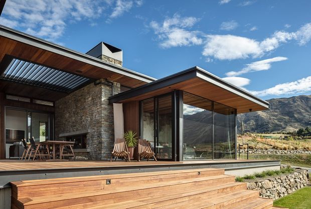 Contemporary mountain home exterior with broad timber decking and steps, a low-pitched projecting roof, extensive floor-to-ceiling glazing, dark metal-framed windows, warm timber soffits, and a prominent rough stone chimney and wall. An outdoor dining area and lounge chairs sit beneath the covered terrace, with rugged mountains visible beyond.
