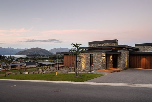 Contemporary single-storey residence viewed from the street at dusk, featuring a low horizontal flat-roof form, extensive natural stone walling, warm timber-clad sections, dark metal roof edges, recessed exterior lighting, and an integrated garage. The landscaped front garden and driveway frame the house, with a mountain and lakeside settlement visible in the background.