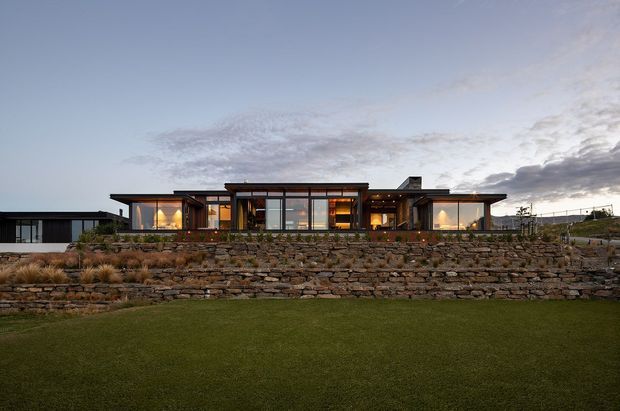 Wide exterior view of a low, single-storey contemporary residence set behind tiered dry-stone retaining walls and a manicured lawn. The home has a long horizontal form, extensive floor-to-ceiling glazing, dark slim frames, timber-lined sections, broad shallow roof planes, and warm interior lighting visible through the windows. The rugged stone landscaping contrasts with the refined modern facade.