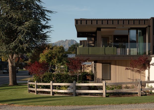 Exterior view of a contemporary two-storey residential house with dark timber-toned cladding, deep roof overhangs, large glazed openings, and a glass-balustraded upper balcony. The house sits within a landscaped suburban setting with mature trees, lawn, a low timber fence, and mountain peaks visible in the distance.