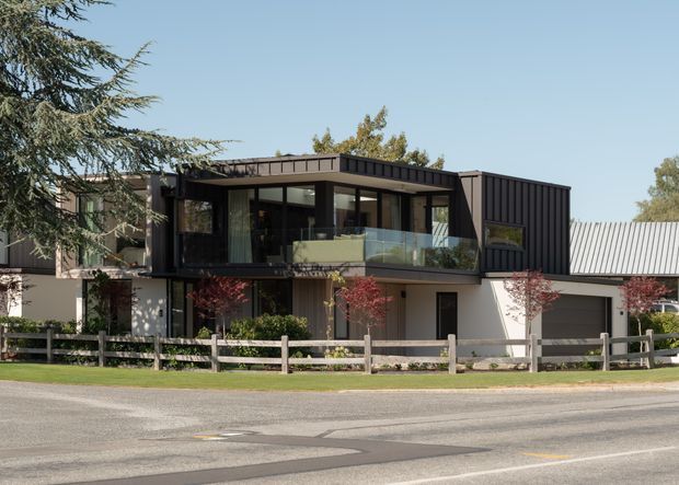 Exterior view of a contemporary two-storey residential building with a dark metal-clad upper level, broad glazing, a glass balcony, white lower-level walls, and an attached garage. The property sits behind a timber post-and-rail fence with landscaped planting and a street in the foreground.