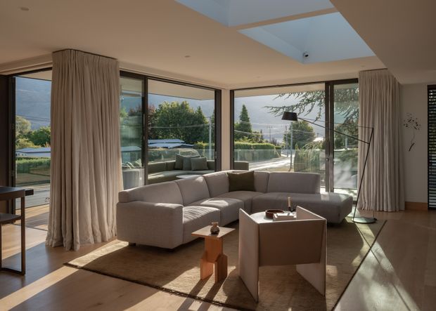A bright, contemporary living room with a large sectional sofa and armchair arranged around a low coffee table. Full-height glazed doors and windows wrap around the room, opening views toward a landscaped garden and distant hills. Floor-to-ceiling curtains, warm timber flooring, a skylight, and restrained neutral furnishings create an airy, refined interior.