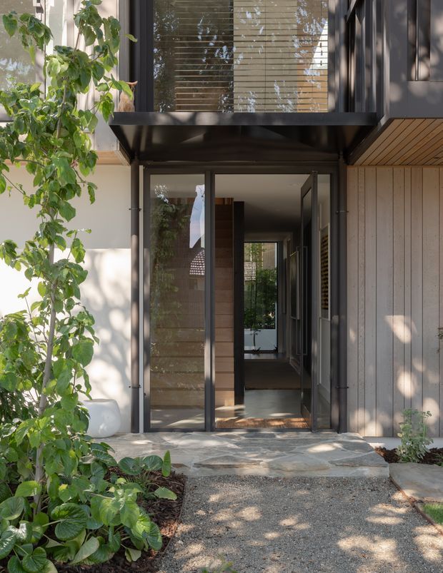 A contemporary residential entry viewed from the exterior, featuring a tall glazed doorway beneath a projecting upper-level window, dark metal framing, vertical timber cladding, and a timber-lined soffit. A paved and gravel pathway leads through a landscaped garden to the recessed entrance. The entry opens toward a bright interior corridor with layered views through the house.