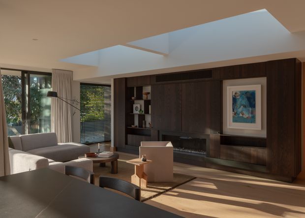 A refined contemporary living room viewed from the dining area, featuring a large dark timber built-in media and fireplace wall, low-profile upholstered seating, a timber coffee table, and expansive glazing with blinds and curtains. A broad ceiling skylight brings strong natural light into the room, creating patterned shadows across the timber floor.