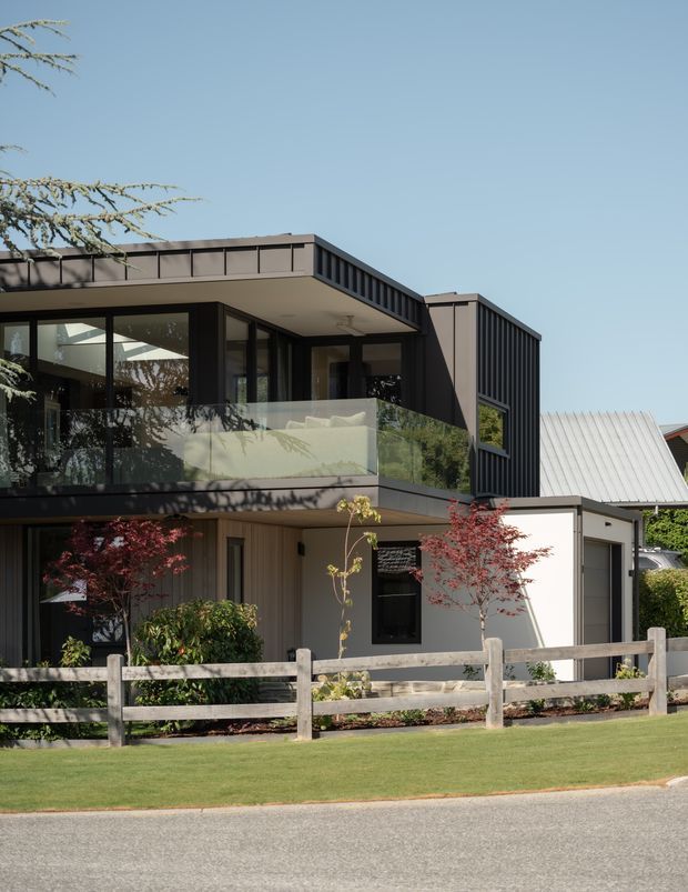 A contemporary multi-level residential house viewed from the street, featuring dark vertical metal cladding, broad flat roof forms, extensive black-framed glazing, and a glass balcony balustrade. A lighter lower-level facade, landscaped front garden, timber post-and-rail fence, and adjacent pitched-roof structure complete the exterior composition.