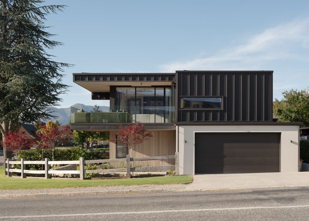 Contemporary two-storey residential house viewed from the street, with a dark vertically ribbed upper facade, projecting flat-roofed living volume, extensive glazing, a timber-toned lower facade, and an integrated double garage. A small landscaped front yard with fencing and planting sits in front of the home, with mountains visible in the distance.