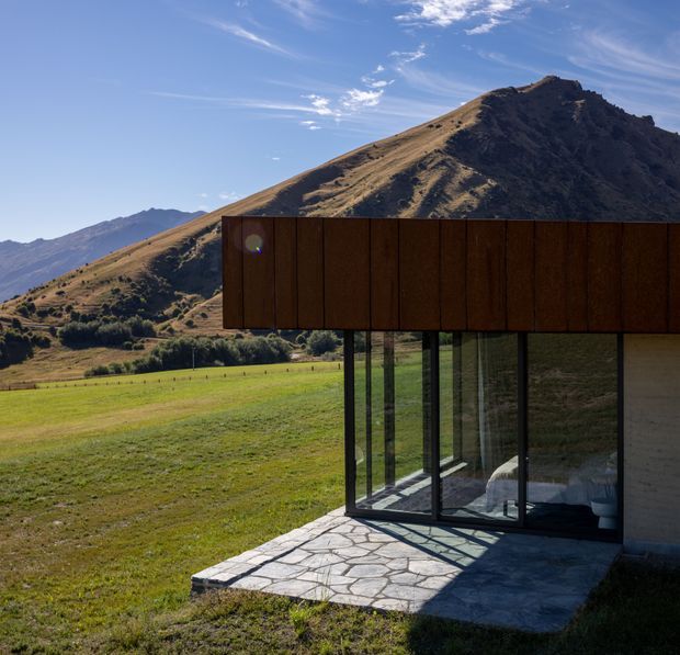 A contemporary single-storey building set in an open alpine or rural landscape, with a projecting dark timber-clad upper volume supported over a fully glazed corner. The glazing reflects and reveals a sparsely furnished interior, while an irregular natural-stone paved terrace sits in front. A steep, rugged mountain dominates the background.